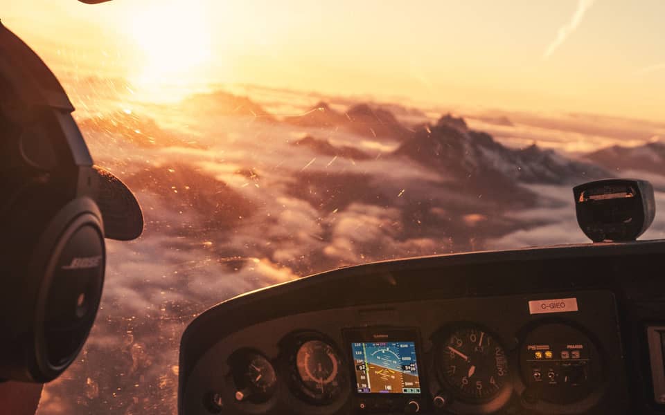 Vedere din scaunul stâng al unui avion ușor, la răsărit: un pilot cu căști privește peste nori și vârfuri de munte, cu bordul dedesubt.